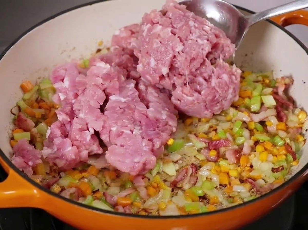 Una cucchiaiata di carne macinata cruda viene aggiunta a una pentola con verdure a cubetti e pancetta in cottura.