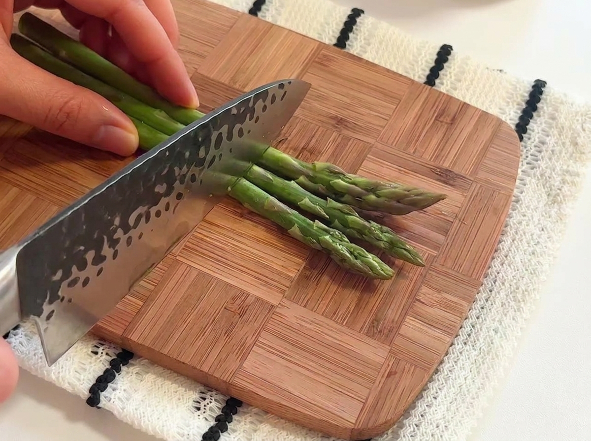 Mani che usano un coltello da chef per tagliare dei gambi di asparagi verdi freschi a pezzetti su un tagliere di legno.