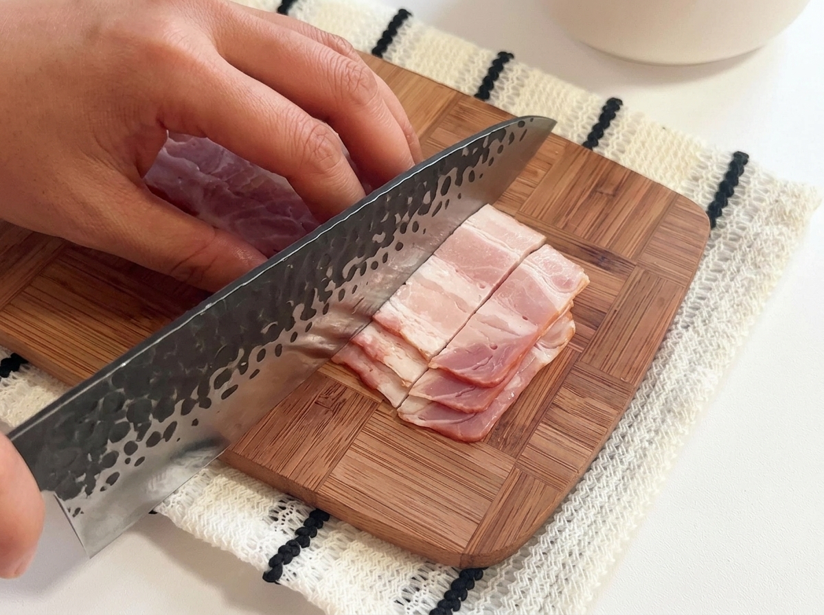 Fette di bacon crudo tagliate a pezzetti su un tagliere di legno con un grosso coltello.