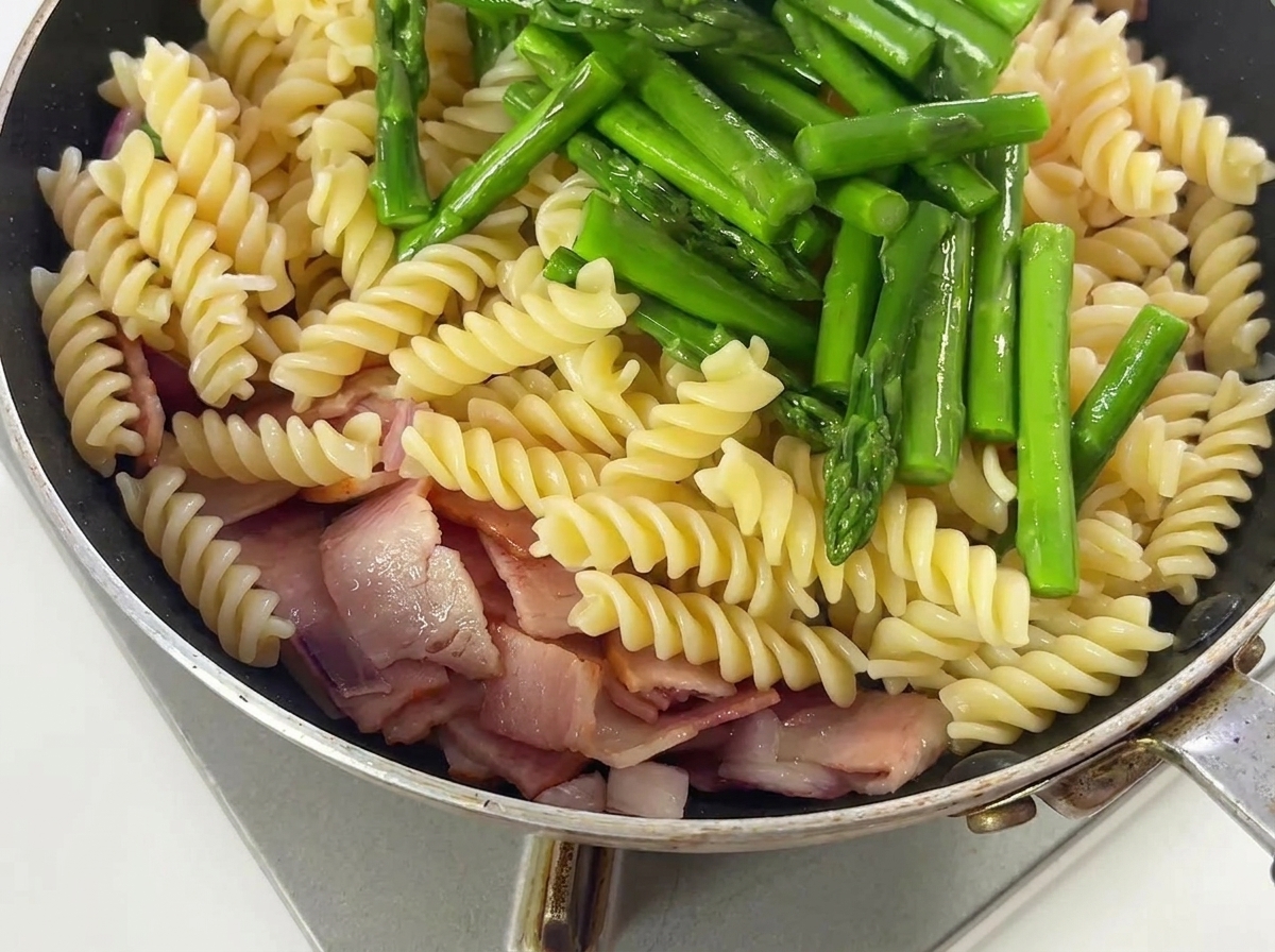 Punte di asparagi e condimenti saltati con pasta, bacon e cipolle in una padella.