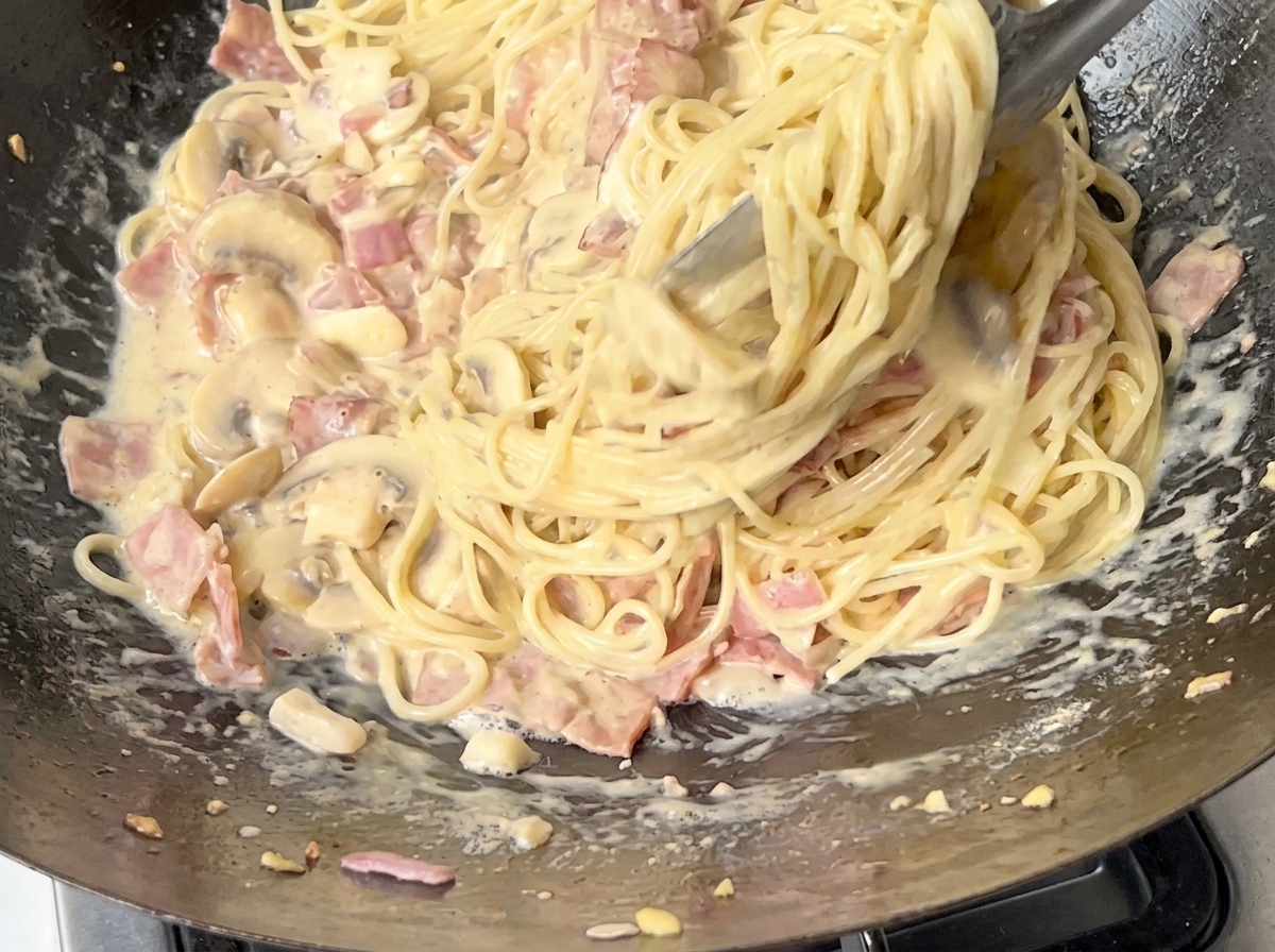 Dettaglio di spaghetti saltati nella salsa cremosa con pinze metalliche.