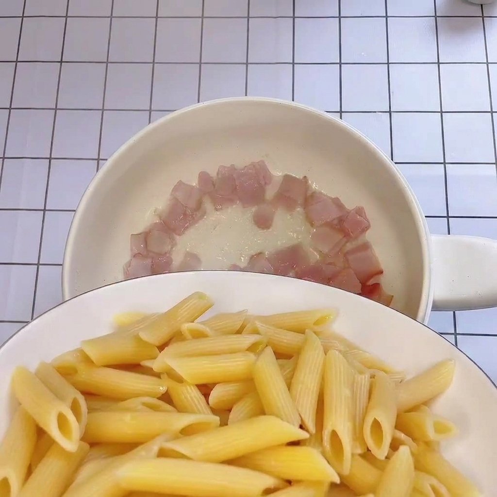 Penne appena scolate aggiunte in una padella contenente pezzi di bacon croccante.