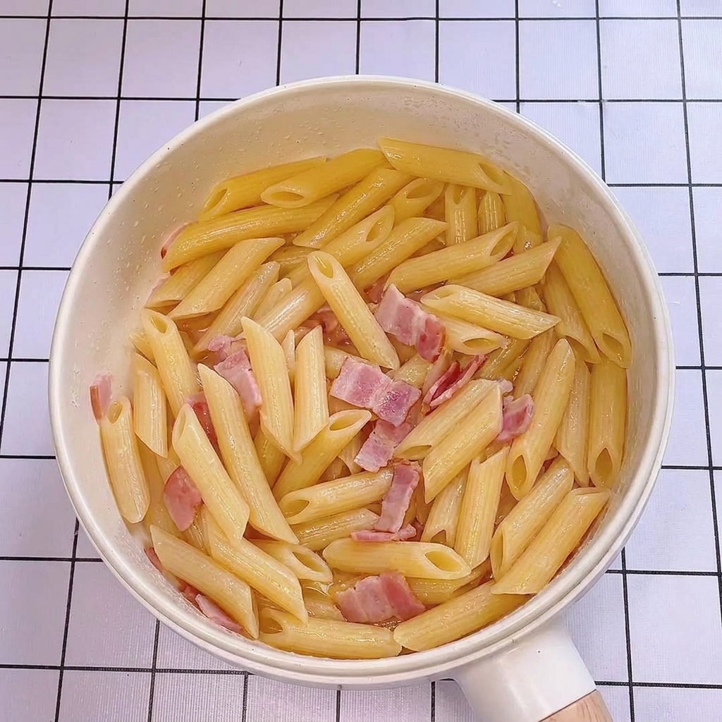 Acqua di cottura della pasta versata nella padella con penne e bacon, creando un aspetto lucido.