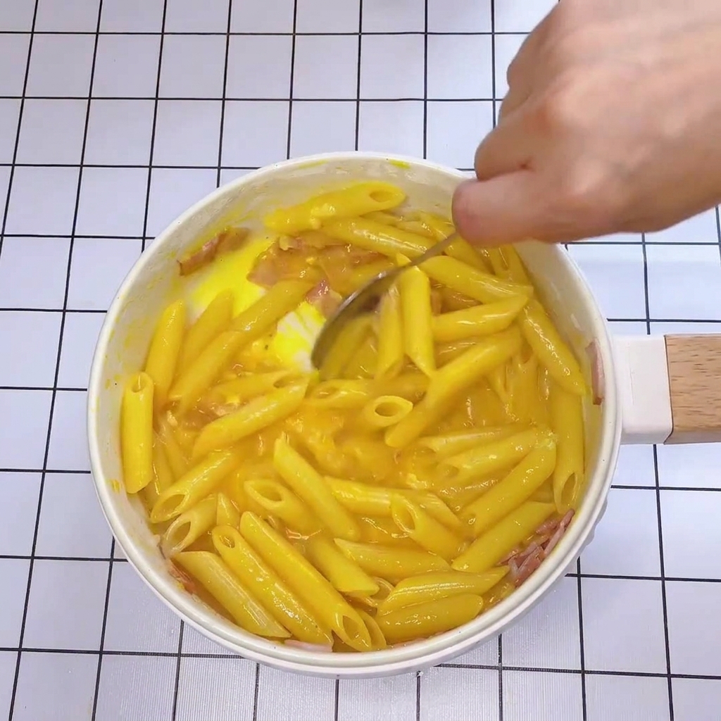Primo piano di un cucchiaio che mescola rapidamente pasta e bacon in padella con una densa salsa gialla.