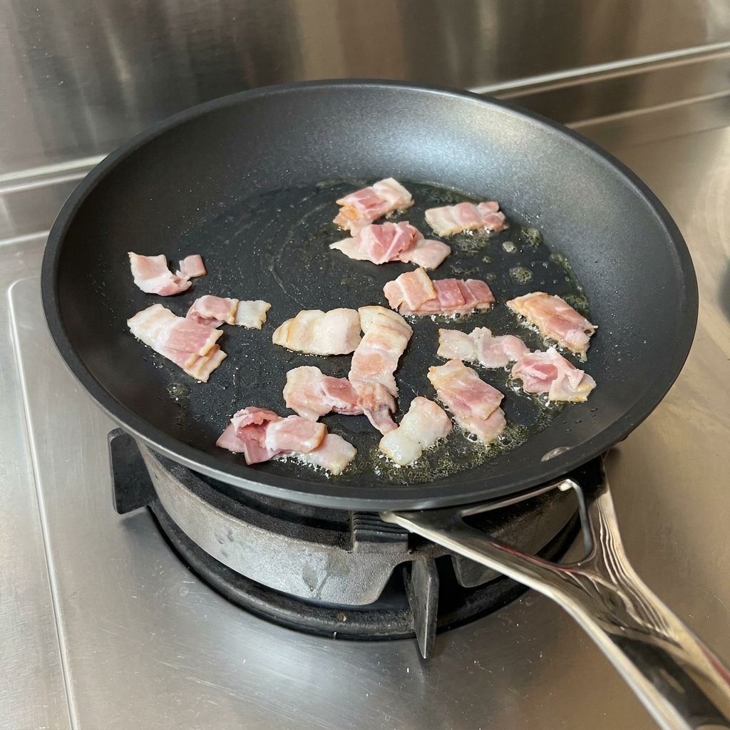 Strisce di pancetta che friggono in padella con olio, mescolate da una spatola rossa.