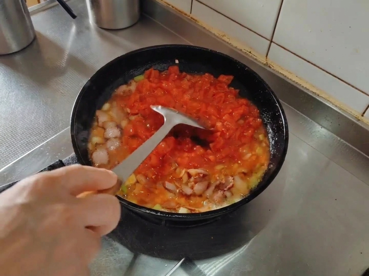 Una spatola di metallo che schiaccia i pomodori in una padella mentre cuociono con pancetta e aromi.
