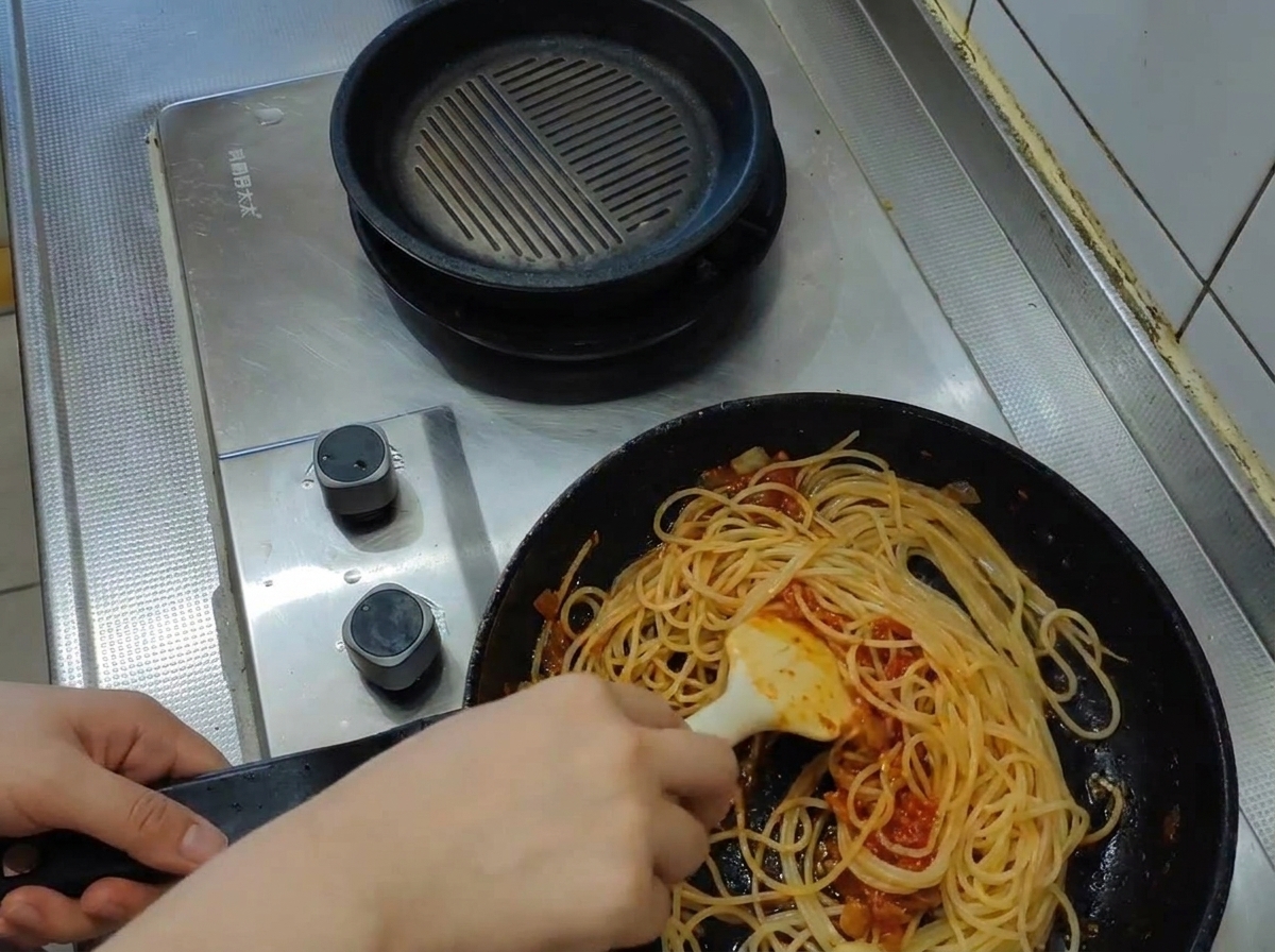 Un mestolo che versa acqua di cottura torbida in una padella con spaghetti e sugo di pomodoro.