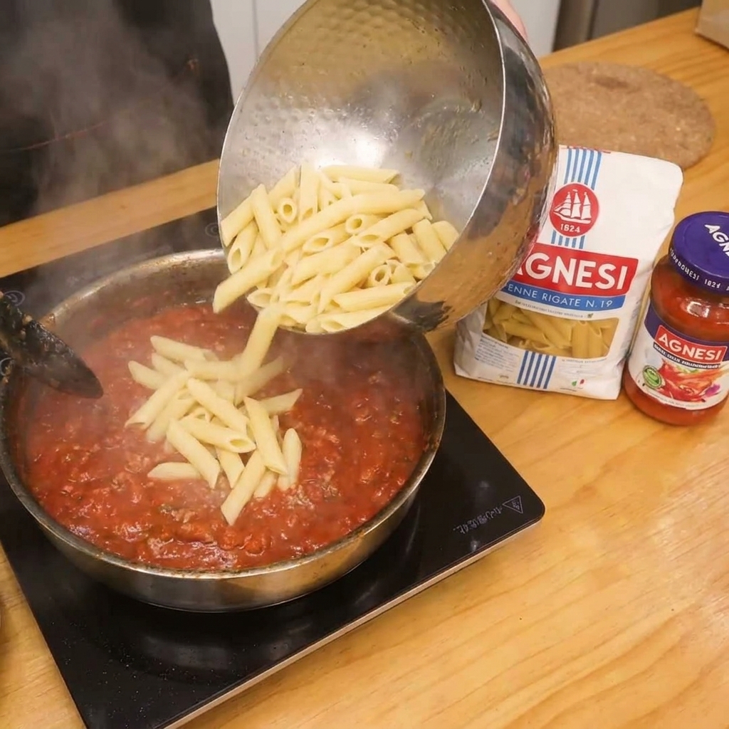 Penne aggiunte al sugo di pomodoro e pancetta nella padella per la mantecatura finale.