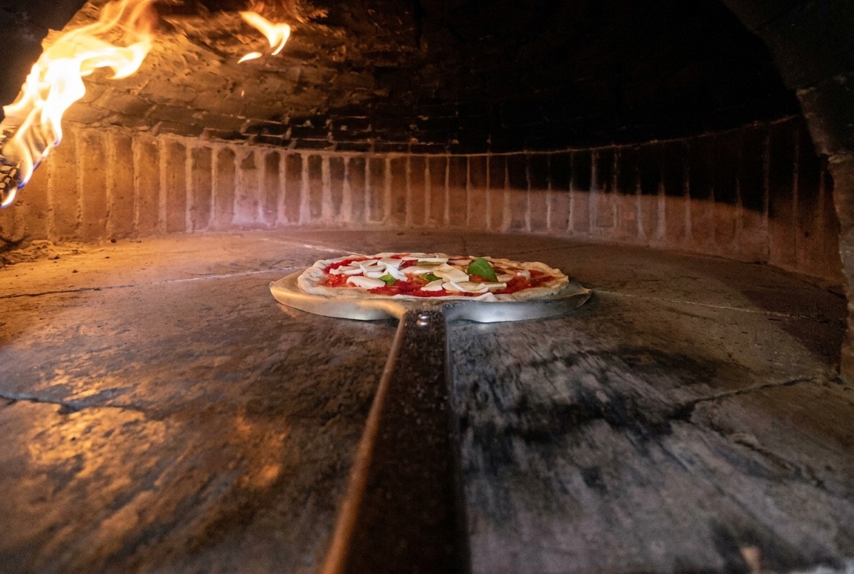 Una pala metallica trasferisce la pizza cruda allinterno di un forno a legna rovente.