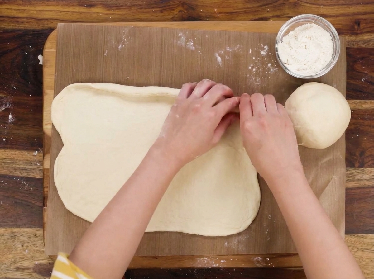 Mani che ripiegano i bordi di un ovale di pasta per formare una barchetta, con una ciotola di formaggio accanto.