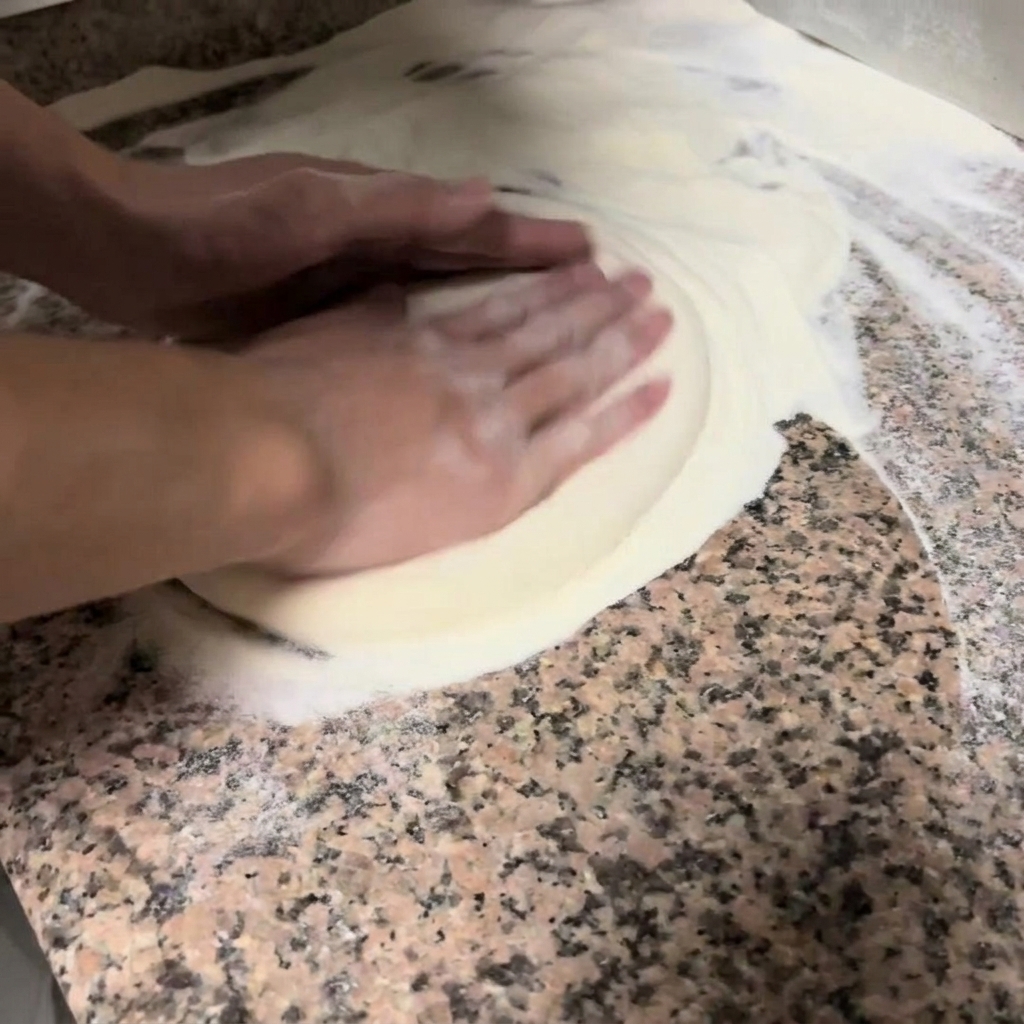 Mani che premono e allargano delicatamente limpasto della pizza su un piano infarinato.