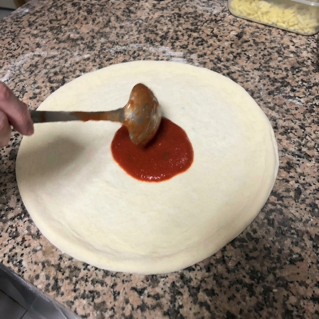 Distribuzione della salsa di pomodoro sulla pizza con un movimento circolare usando un mestolo di metallo.