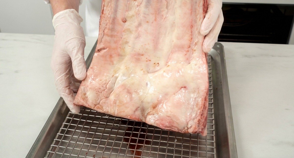Un grande pezzo di costine di manzo crude con venature di grasso bianco, tenuto da uno chef sopra una griglia metallica.