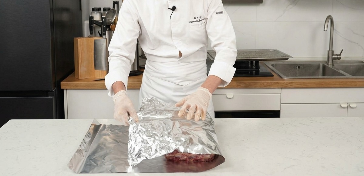 Uno chef che piega un grande foglio di alluminio sopra le costine di manzo condite su un piano da cucina.