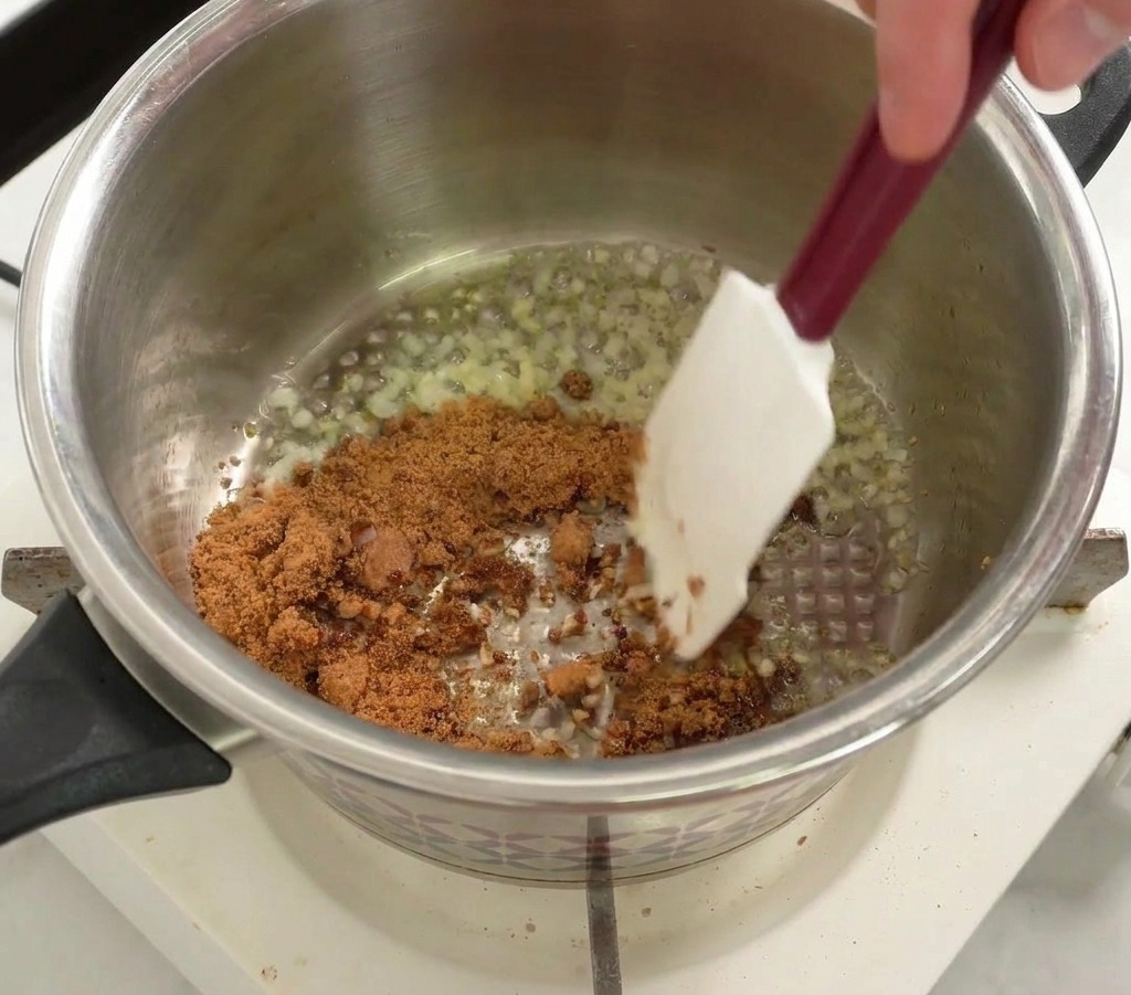 Zucchero di canna mescolato in una pentola con cipolla e aglio soffritti usando una spatola bianca.