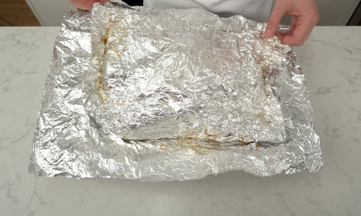 Mani che aprono con cura lalluminio per rivelare un grande pezzo di costine di manzo cotte lentamente su un piano da cucina.