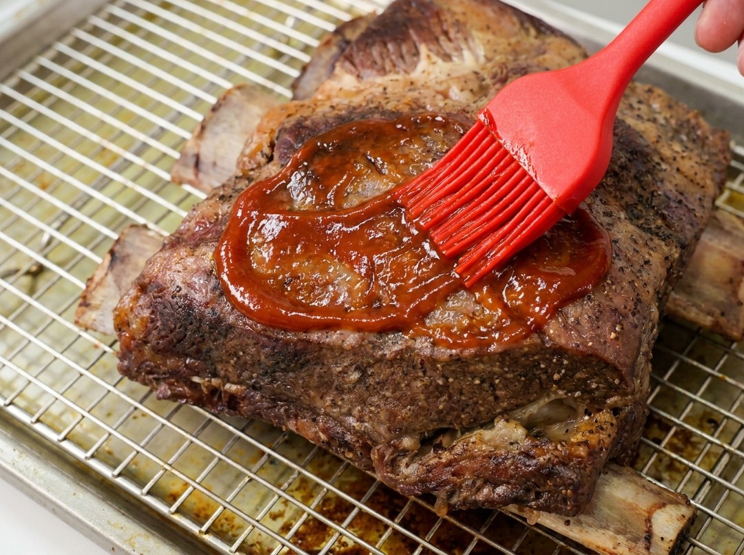 Una mano che usa un pennello in silicone rosso per spalmare una densa salsa BBQ lucida su costine di manzo su una griglia.