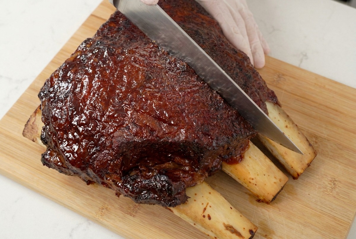 Un coltello da chef che taglia costine di manzo scure e caramellate su un tagliere di legno, mostrando la carne succosa.