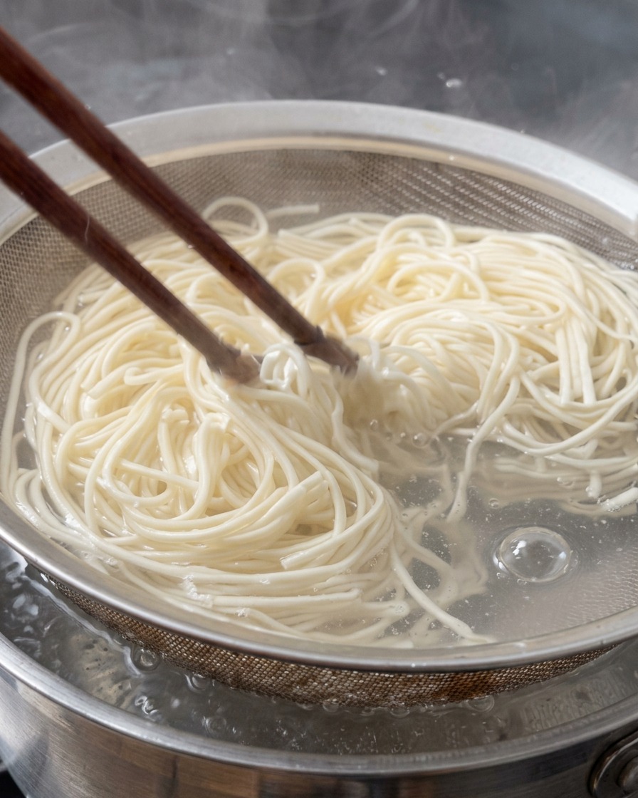 Noodles ramen freschi sollevati dallacqua bollente con un colino a maglia metallica e bacchette di legno.
