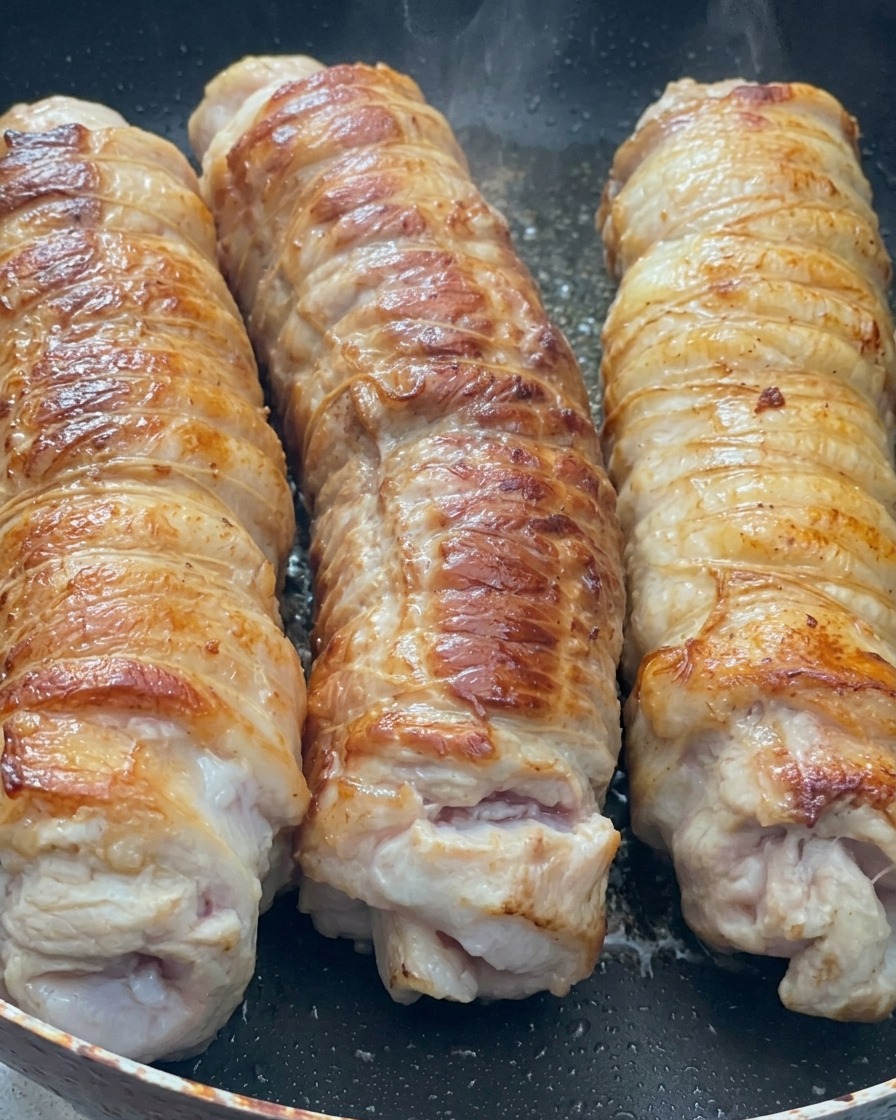 Tre rotoli di pancetta di maiale che rosolano in una padella, mostrando una crosta dorata e caramellata.