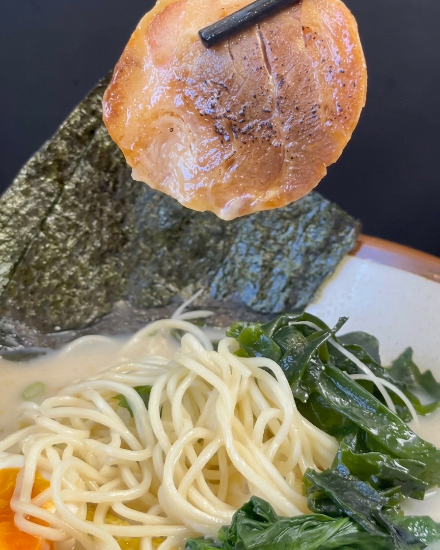 Bacchette nere che sollevano una fetta di maiale chashu sopra una ciotola di ramen assemblata.