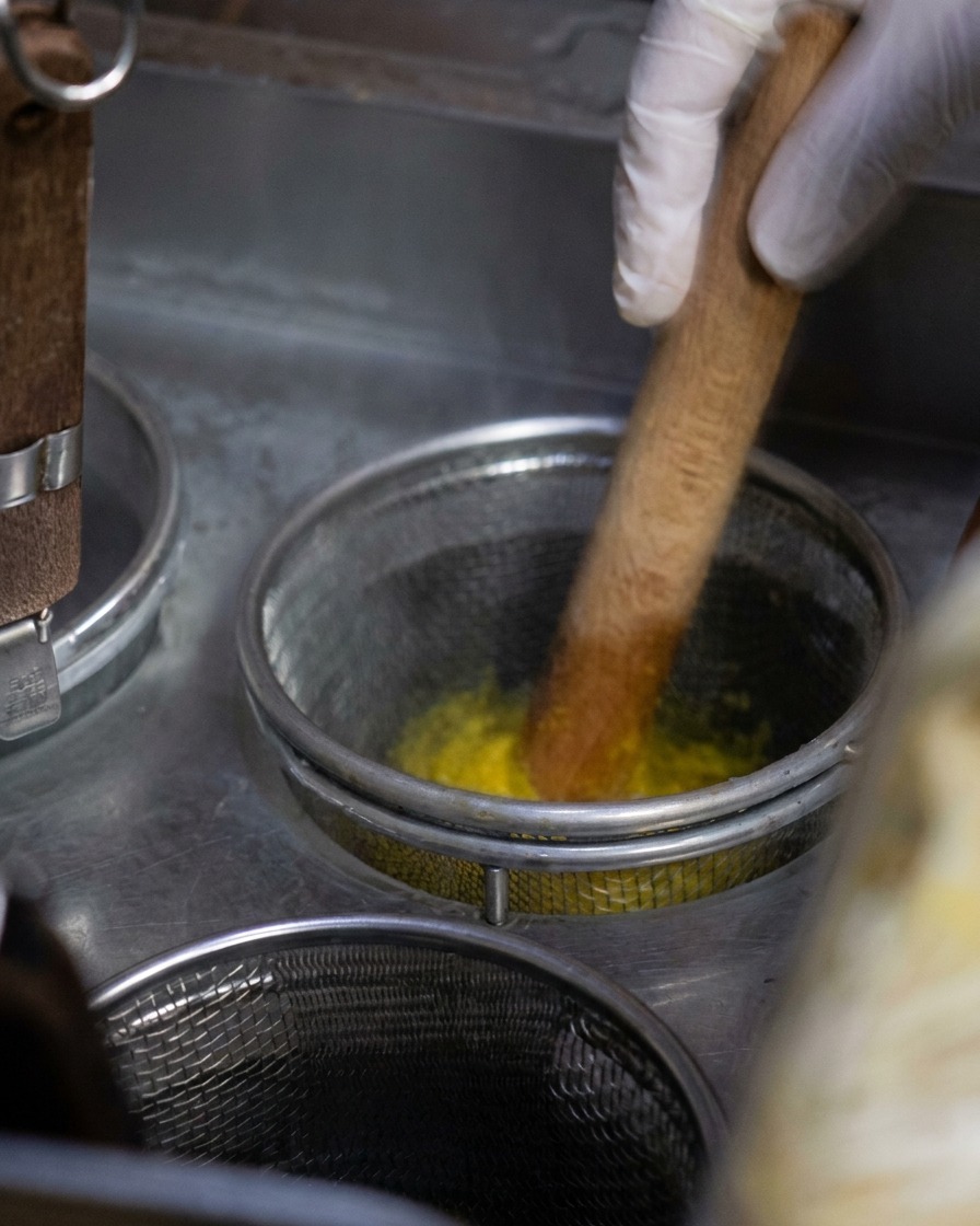 Un cestello in rete metallica pieno di noodles gialli mentre vengono mescolati con un utensile in legno in una pentola di acqua bollente.
