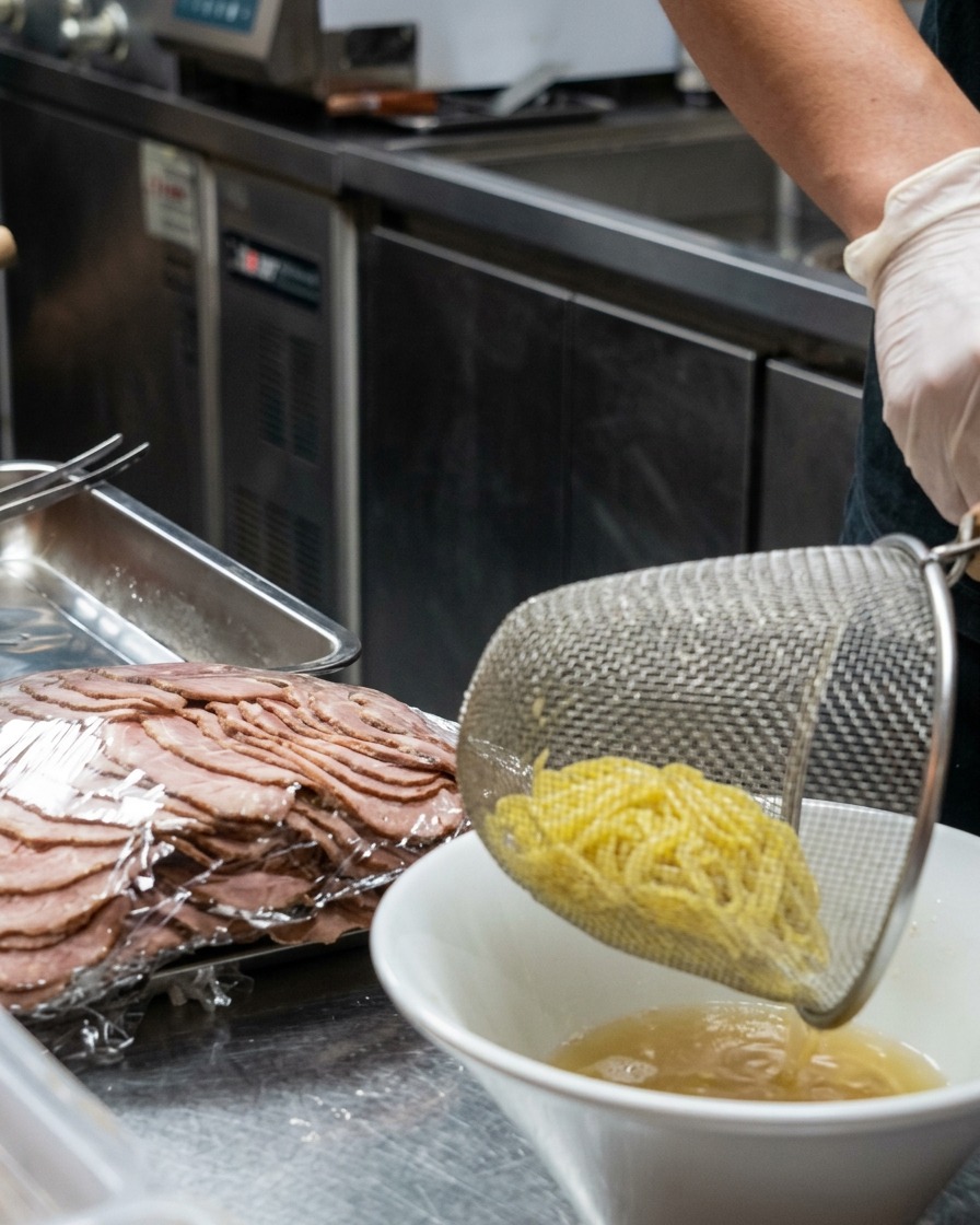 Un colino in metallo che lascia cadere noodles gialli appena cotti in una ciotola bianca piena di brodo cremoso.