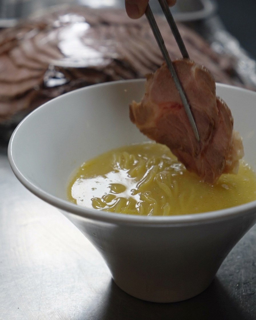 Bacchette di metallo che posano una fetta spessa di maiale scottato in una ciotola di ramen.