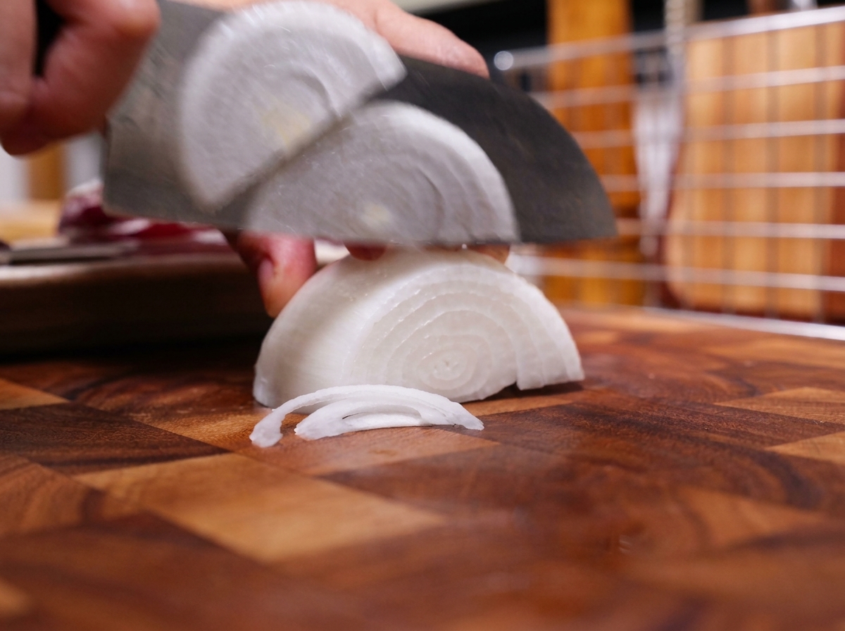 Un coltello da chef affetta una metà di cipolla bianca in strisce sottili su un tagliere di legno.