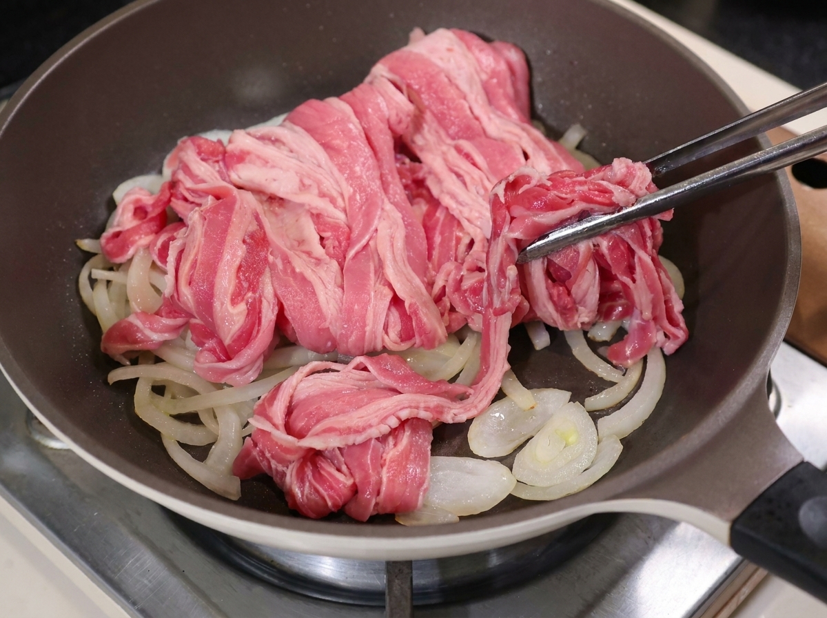 Pinze in metallo posizionano fettine di manzo crudo sopra le cipolle cotte in padella.