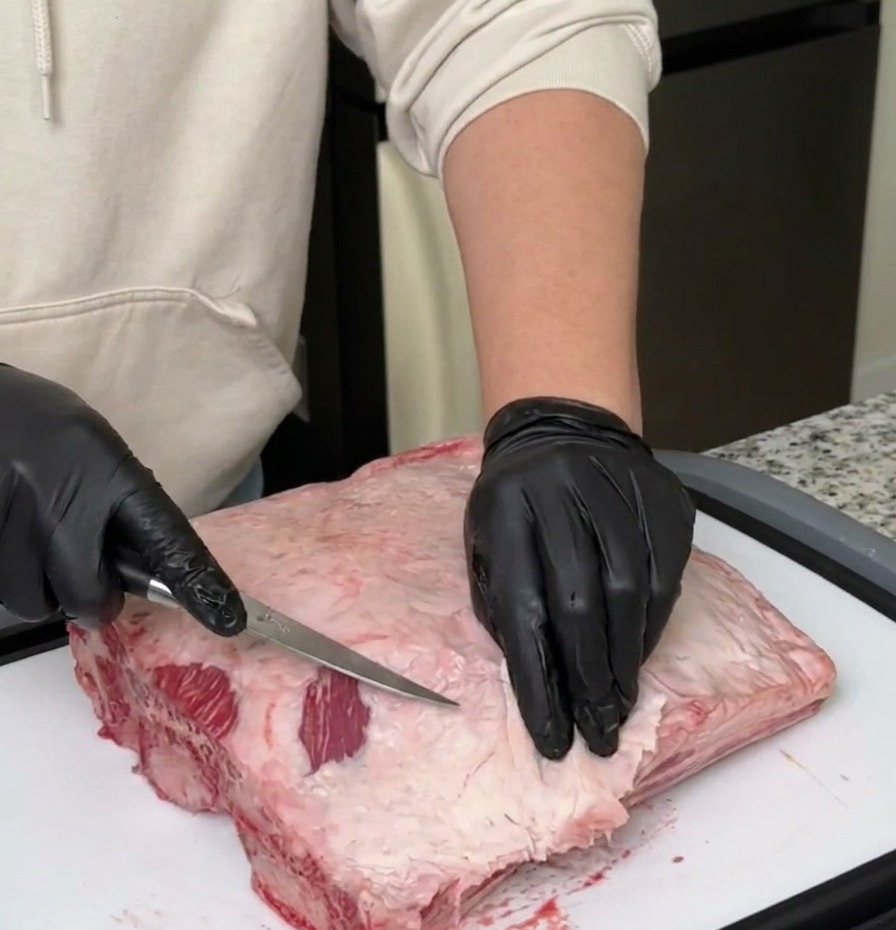 Una persona con guanti neri usa un coltello da disosso affilato per rimuovere grasso e membrana da un pezzo di costine di manzo crude.