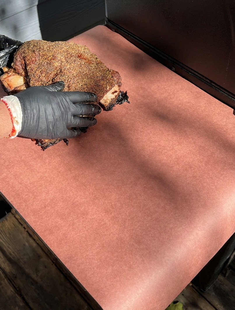 Mani guantate posizionano le costine di manzo affumicate su un grande foglio di carta da macellaio rosa per avvolgerle.