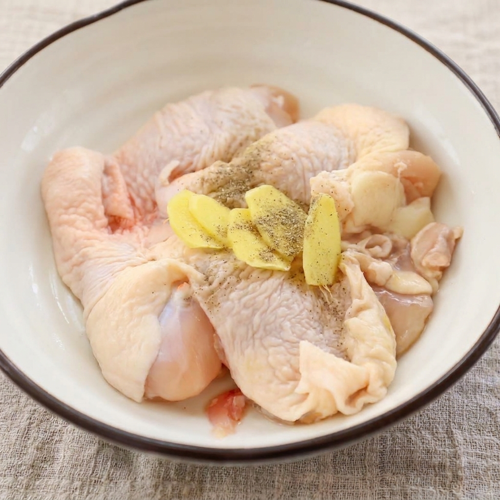 Cosce di pollo crude in una ciotola, guarnite con fette di zenzero fresco e pepe nero macinato.