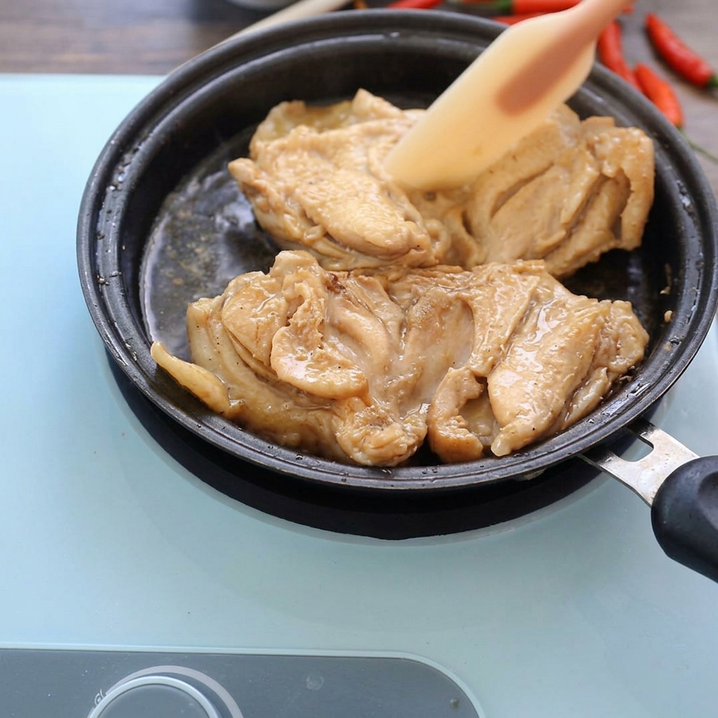Cosce di pollo cotte che sobbollono in una padella bassa con una salsa scura, premute leggermente con una spatola in silicone.