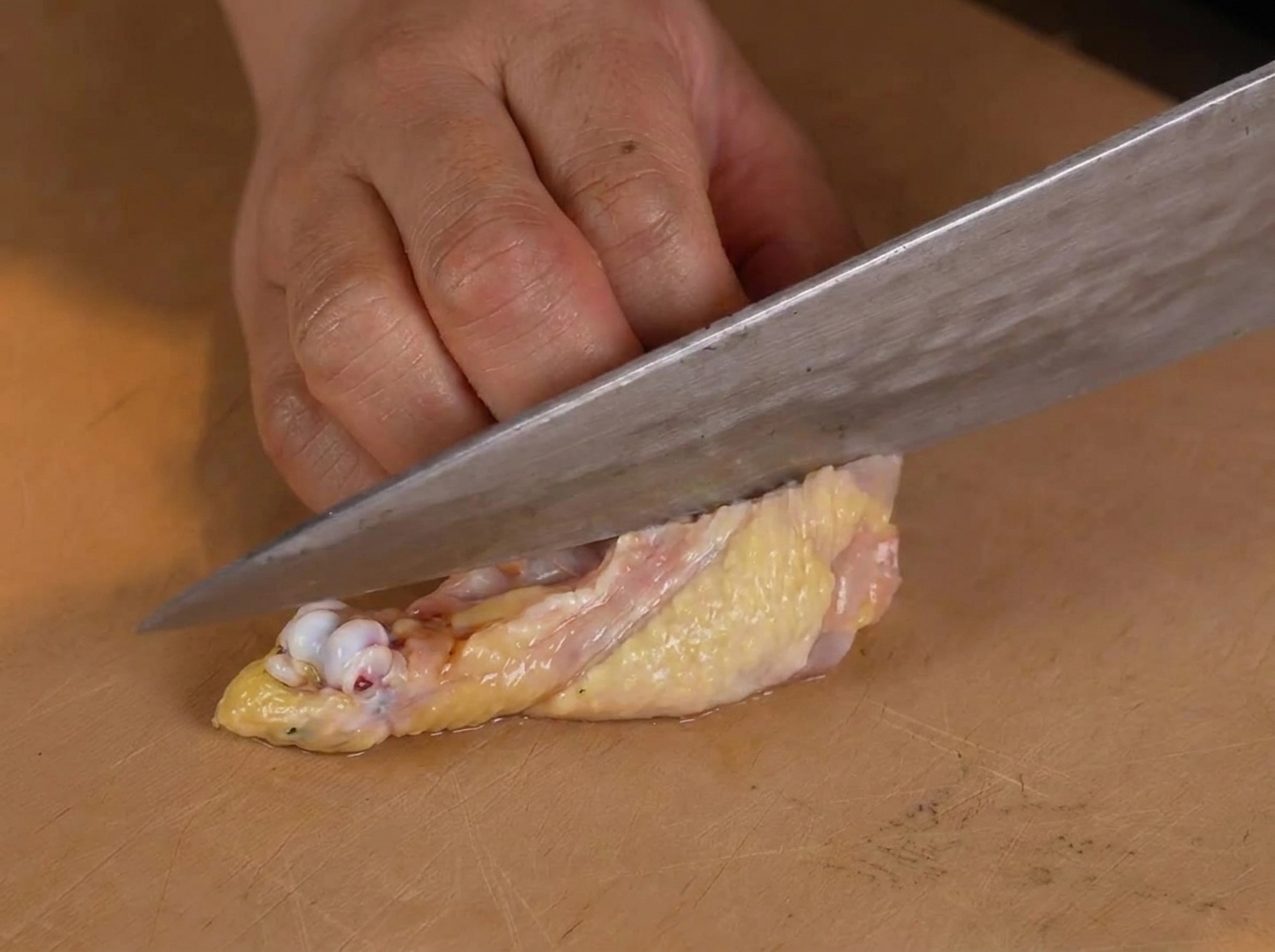 Una persona usa un coltello da chef per rimuovere con cura losso da una coscia di pollo cruda su un tagliere di legno.