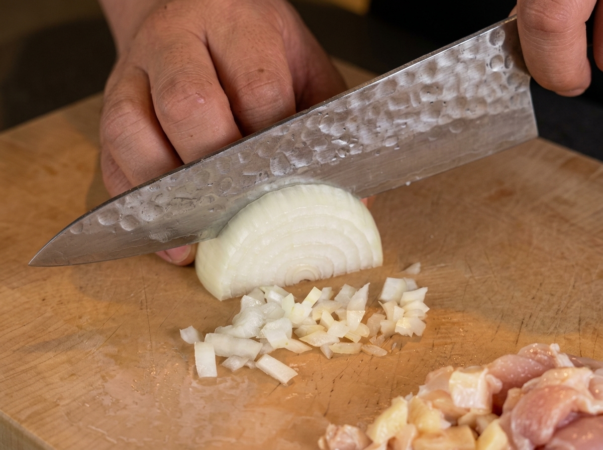 La mano di uno chef usa un coltello per tritare finemente mezza cipolla bianca su un tagliere di legno.