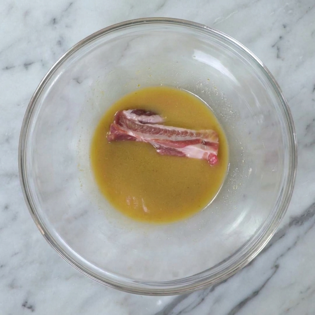 Una singola costina di maiale cruda immersa nella marinata di miso dorata allinterno di una ciotola di vetro.