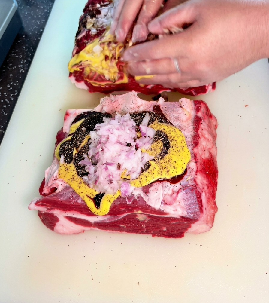Mani che massaggiano attivamente un mix di cipolle tritate, senape e salse scure su grandi fette di carne cruda.