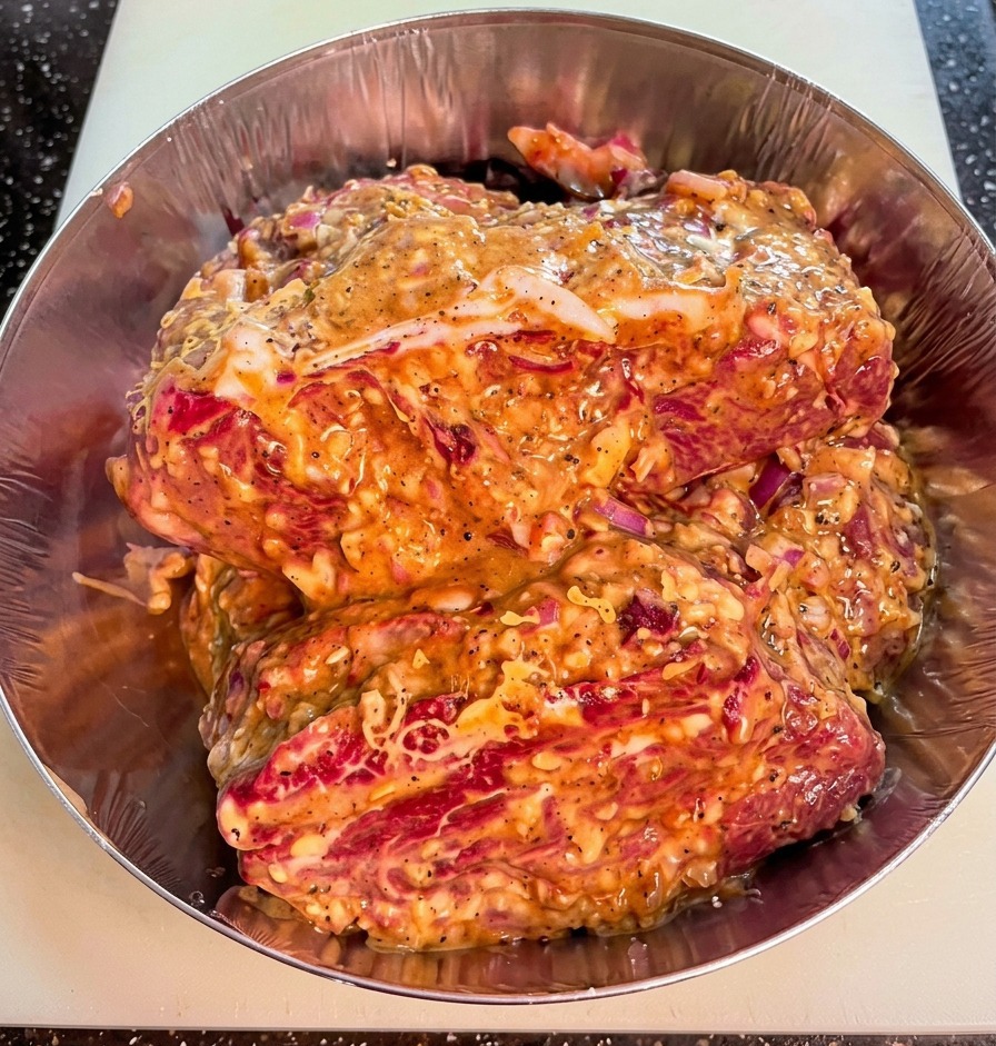 Primo piano di pezzi di manzo crudo in una ciotola di metallo, ricoperti da una marinata densa con pezzi visibili di cipolla e pepe.