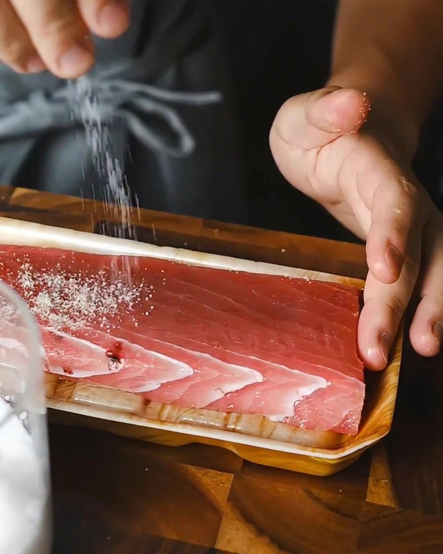 Una persona che cosparge uniformemente del sale su un blocco rettangolare di tonno crudo su un vassoio.