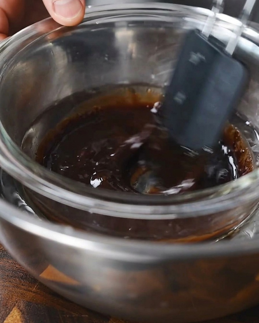 Una spatola che mescola una marinata liquida scura in una ciotola di vetro posta su un bagno maria.