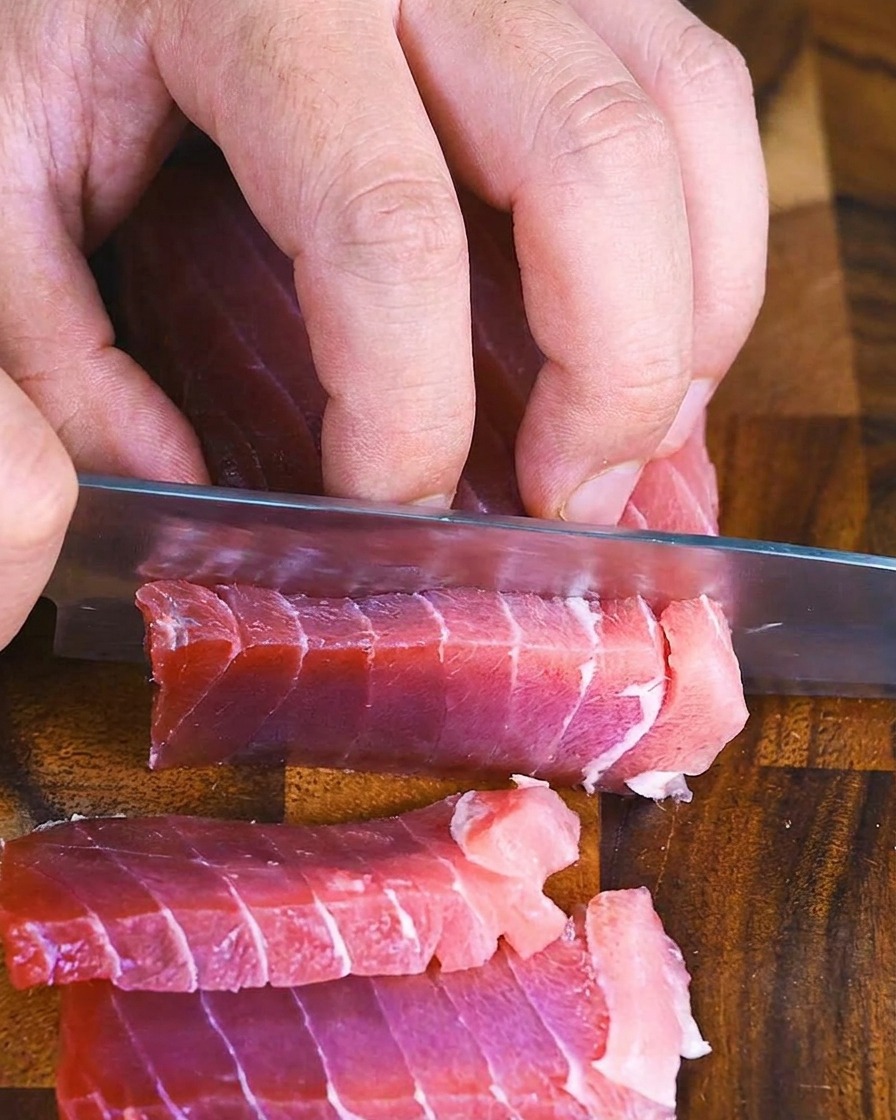 Mani che usano un coltello da chef per affettare un blocco di tonno crudo in rettangoli spessi e uniformi su un tagliere di legno.