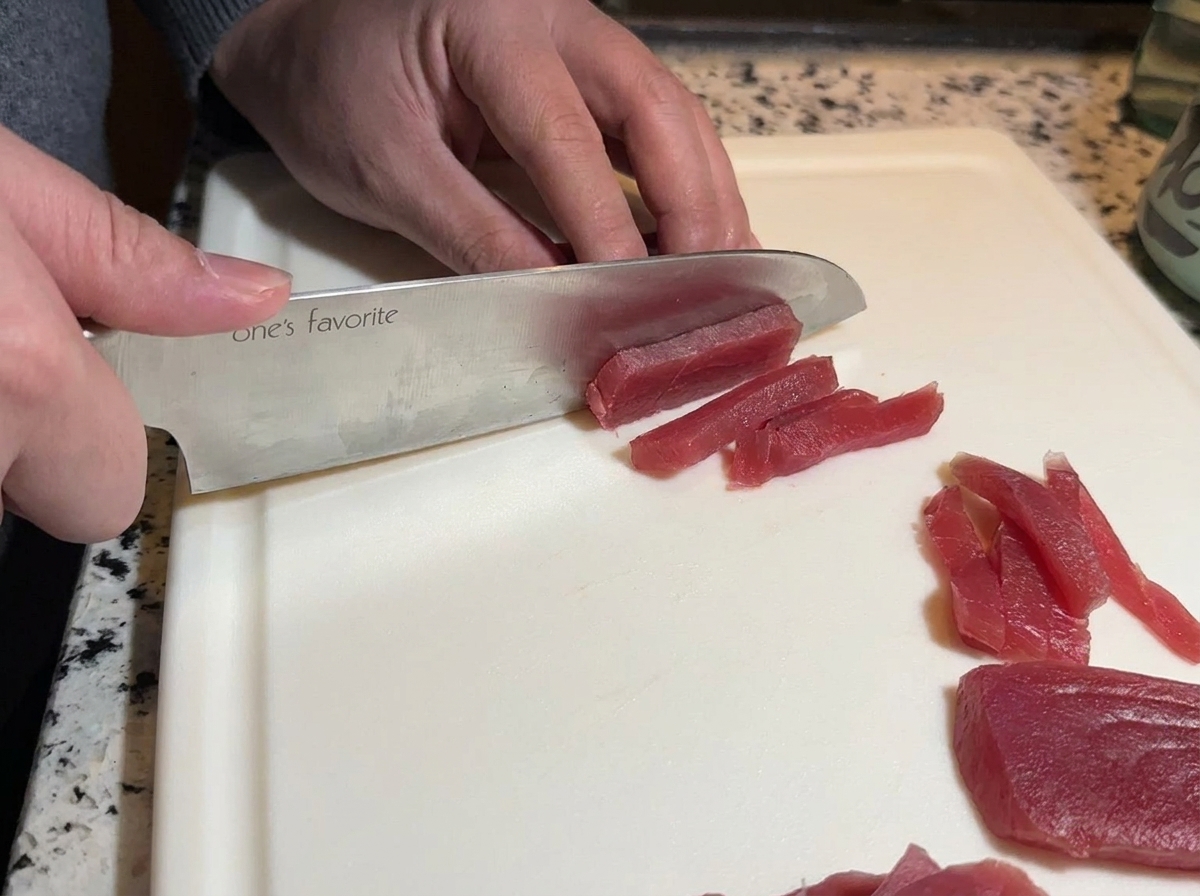 Un coltello da chef affilato che taglia un pezzo di tonno rosso crudo in strisce lunghe su un tagliere bianco.