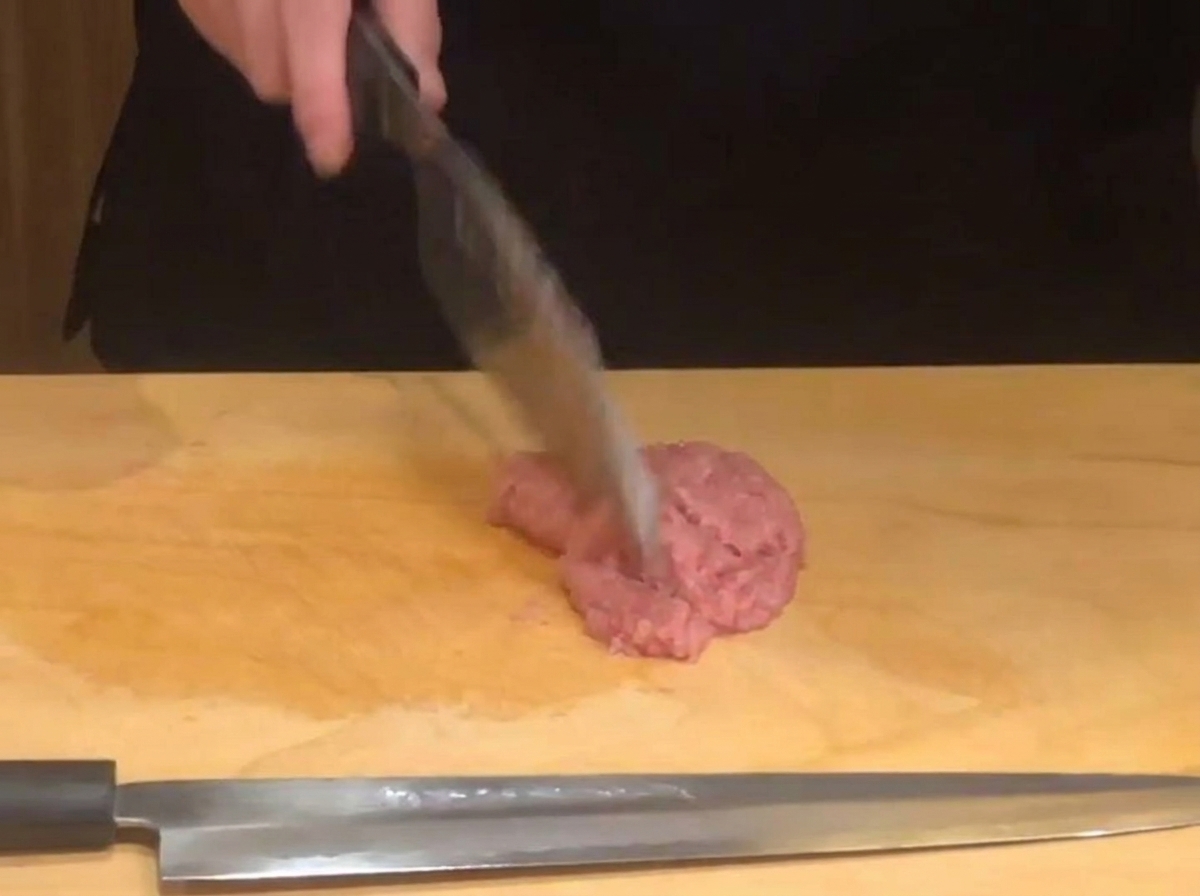 Mano di uno chef che usa un coltello affilato per tritare il tonno crudo rosa su un tagliere di legno.