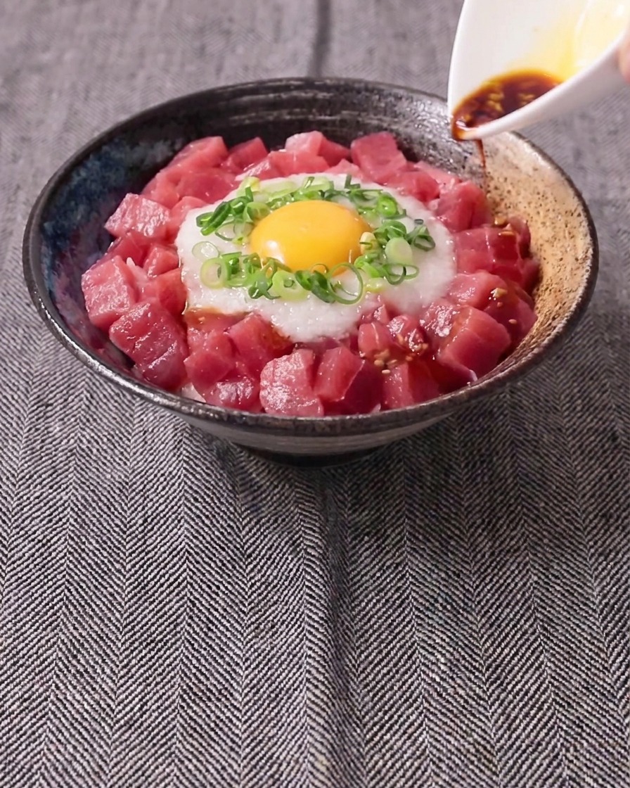 Una salsa scura versata da una piccola brocca bianca su una ciotola di tonno crudo a cubetti, igname bianco grattugiato, cipollotto e un tuorlo duovo crudo.