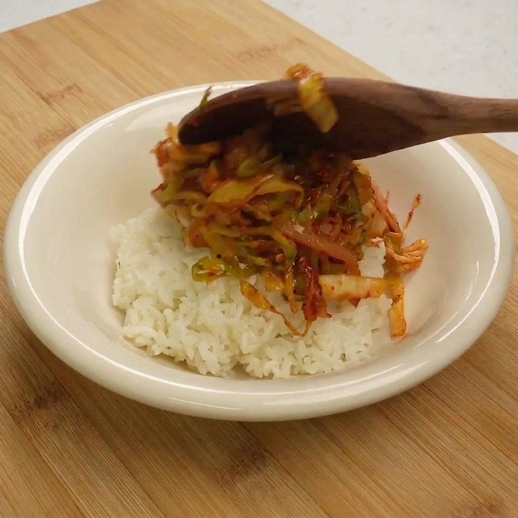 Un cucchiaio di legno che serve lo stir-fry di tonno e cavolo sopra una porzione di riso bianco in una ciotola.