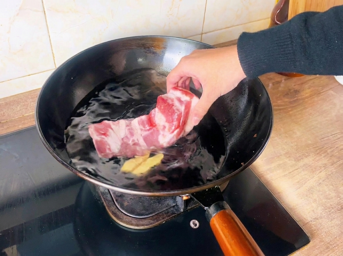 Una mano che mette un pezzo di pancia di maiale cruda in un wok pieno di acqua fredda e fette di zenzero.