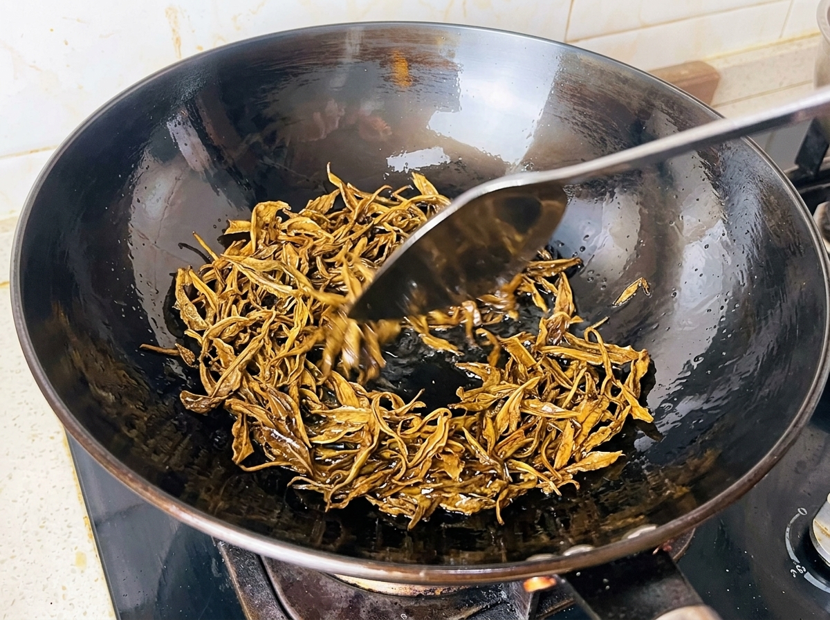 Foglie di tè verde bagnate fritte in olio caldo allinterno di un wok nero con una spatola di metallo.