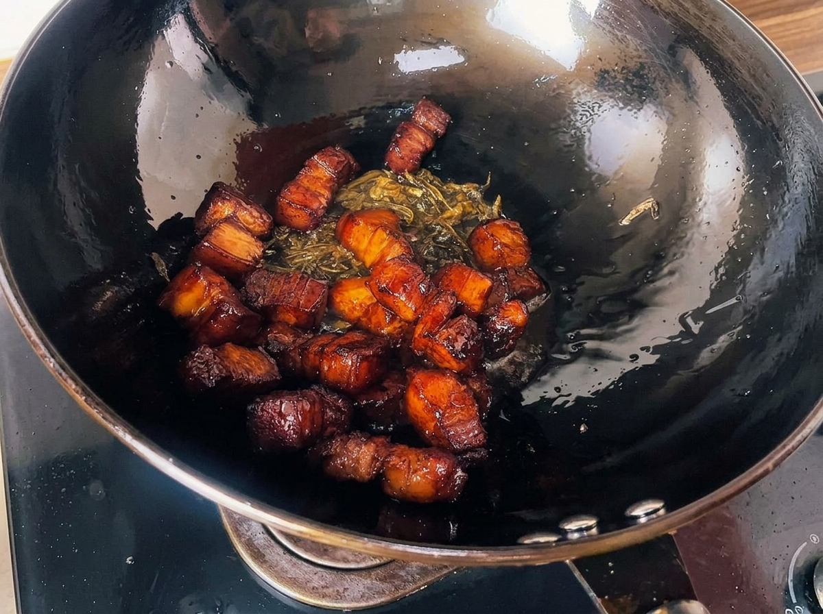 Cubetti di pancia di maiale arrostiti e glassati mescolati con foglie di tè croccanti allinterno di un grande wok nero.