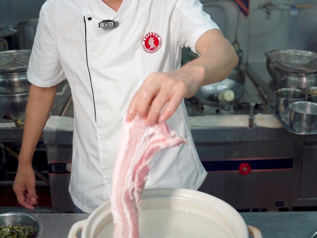 Uno chef che tiene un grosso pezzo di pancetta cruda sopra una pentola bianca piena dacqua.